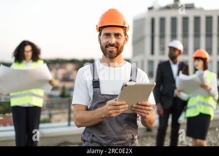 Portrait de contremaître barbu souriant en casque dur et uniforme posant sur la caméra avec tablette numérique dans les mains. Équipe d'investisseurs multiethniques vérifiant le projet de construction tout en se tenant sur la terrasse panoramique. Banque D'Images