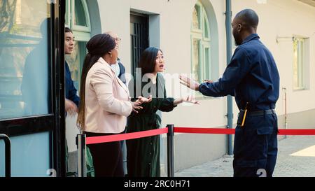 Garde de sécurité demandant aux gens d'attendre, essayant de raisonner avec une foule de clients anxieux attendant depuis les premières heures du matin. Diverses personnes assises à l'extérieur du magasin de vêtements, promotions du vendredi noir. Banque D'Images