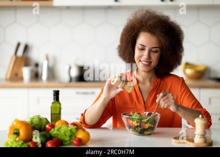 Heureuse jeune dame noire préparant une salade de légumes saine dans la cuisine, ajoutant du sel Banque D'Images