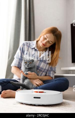 Un aspirateur robot intelligent nettoie le tapis, une fille et un chat regardent l'aspirateur. Le concept de maison intelligente, nettoyage quotidien Banque D'Images