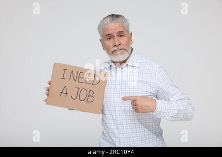 Homme senior sans emploi pointant du doigt le signe en carton avec la phrase I Need A Job sur fond blanc Banque D'Images