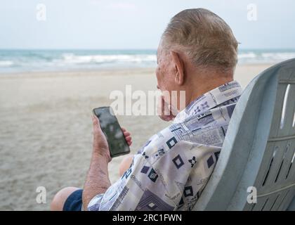 Homme âgé assis sur une chaise de plage et utilisant son smartphone pour lire. Utilisation de la technologie pour les personnes âgées. Banque D'Images