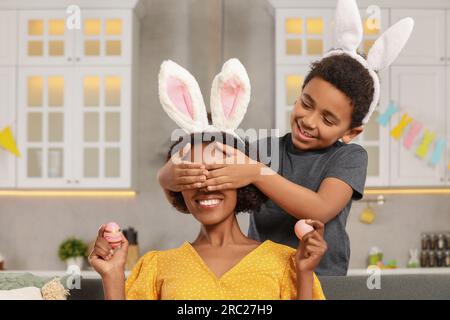 Mignon fils afro-américain couvrant les yeux de sa mère avec les mains alors qu'elle tient des oeufs de Pâques dans la cuisine Banque D'Images