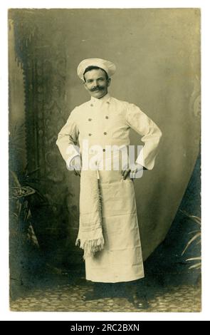 Carte postale originale du début des années 1900 d'un chef masculin joyeux, moustache, en uniforme blanc portant un chapeau de chef, serviette en tablier à la ceinture, circa 1910, Royaume-Uni Banque D'Images