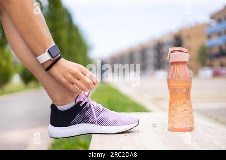 Femme sportive attachant des lacets sur un banc à l'entraînement de fitness en plein air. Entraînement de course à pied pour jeune athlète féminine. Hydratation, bien-être, exercice, santé. Banque D'Images