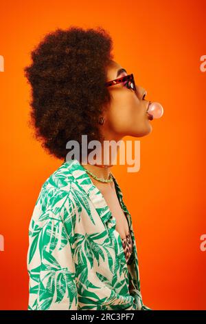 Vue de côté de jeune et tendance femme afro-américaine dans des lunettes de soleil portant la tenue d'été tout en soufflant du bubble gum et debout isolé sur rouge, femme Banque D'Images