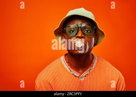 Portrait de jeune et élégant homme afro-américain en tenue d'été portant des lunettes de soleil et chapeau panama soufflant du bubble gum et regardant l'isolat de caméra Banque D'Images