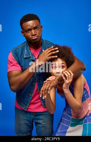 Sérieux jeune homme afro-américain en tenue d'été couvrant le meilleur ami effrayé en robe de robe élégante et regardant la caméra isolée sur bleu, fashionabl Banque D'Images