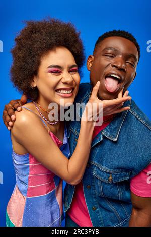 Portrait de jeune femme afro-américaine souriante avec le maquillage audacieux touchant le meilleur ami à la mode sortant de la langue et debout isolé sur bleu, fashi Banque D'Images