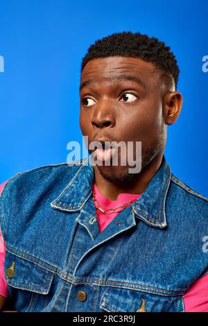 Portrait de jeune homme afro-américain choqué avec coiffure moderne portant la tenue d'été et regardant loin tout en se tenant debout et posant isolé sur le bleu Banque D'Images