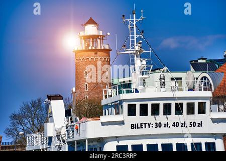 Kolberg, Pologne, 20 avril 2023 : bateau d'excursion devant le phare dans le contre-jour du soleil dans le port de la ville polonaise de Kołobrzeg Banque D'Images