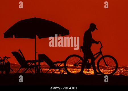 Île de Palms, États-Unis. 12 juillet 2023. Un travailleur de la station silhouette par le lever du soleil pousse un vélo le long de la plage après avoir installé des chaises de plage et un parasol au Wild Dunes Resort, le 12 juillet 2023 à Isle of Palms, Caroline du Sud. Une vague de chaleur prolongée dans le sud des États-Unis continue d'apporter un temps extrêmement chaud et humide à la région. Crédit : Richard Ellis/Richard Ellis/Alamy Live News Banque D'Images