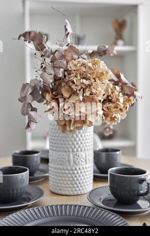 Jolies fleurs dans le vase sur une table à manger avec café ou thé servi tasses et assiettes est prêt pour la réception de quêtes ou le petit déjeuner en famille, gros plan, Banque D'Images