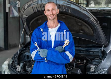 Un mécanicien automobile masculin heureux en combinaison bleue tenant un wranch dans chaque main et croisant ses bras en se tenant devant une voiture avec son capot ouvert Banque D'Images