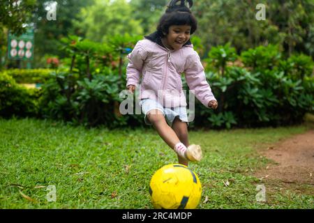 Une jeune fille en uniforme de soccer dribble un ballon de soccer dans un parc. Elle est souriante et semble concentrée sur la tâche à accomplir. Le fond est luxuriant Banque D'Images