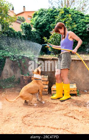 Vue latérale de jeune femme dans des vêtements décontractés et bottes de pluie jaunes debout avec chien joueur obéissant dans la campagne tout en arrosant le jardin en plein jour Banque D'Images