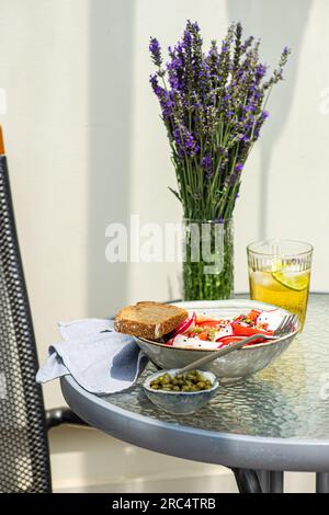 Salade de légumes avec tomates bio et câpres servi dans un bol avec fourchette et tranche de pain près d'un verre de limonade et bol de câpres sur verre extérieur Banque D'Images