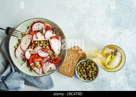 Vue de dessus de la salade de légumes avec tomates bio et câpres servi dans un bol avec fourchette près des tranches de pain, verre de limonade et bol de câpres sur outd Banque D'Images