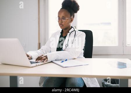 Médecin féminin noir occupé dans des lunettes avec stéthoscope tapant sur l'ordinateur tout en étant assis à table dans la clinique moderne Banque D'Images