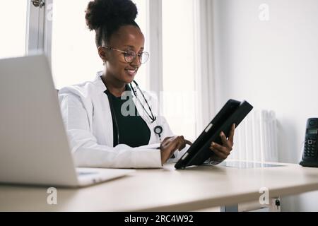 Sourire afro-américaine médecin dans les lunettes, uniforme et stéthoscope regardant l'écran de la tablette et l'information de navigation avec le doigt tandis que s Banque D'Images