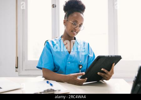 Positif afro-américaine médecin en uniforme bleu et lunettes souriant et regardant l'écran de comprimé dans les mains tout en étant assis à la table avec la Banque D'Images