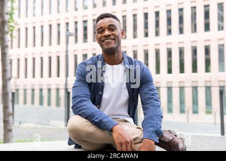 Portrait de amical homme afro-américain dans des vêtements décontractés souriant et regardant la caméra tout en se relaxant sur le banc sur la rue de la ville Banque D'Images
