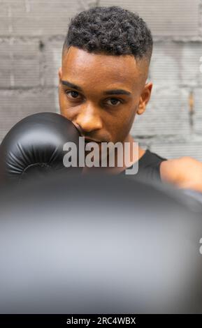 Portrait de boxeur masculin afro-américain en vêtements de sport et gants en cuir pratiquant le punch et regardant la caméra contre le mur de briques dans la salle de gym Banque D'Images