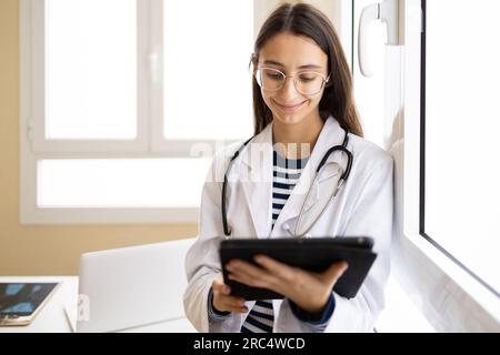 Jeune femme médecin en manteau blanc et lunettes regardant l'écran de tablette tout en se tenant debout dans le bureau de la clinique moderne Banque D'Images