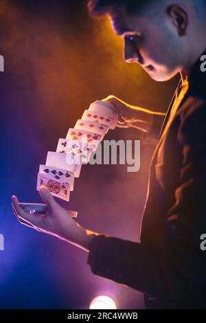 Illusionniste masculin de vue latérale en costume mélanger un paquet de cartes tout en jouant sur scène dans le spectacle contre le mur sombre Banque D'Images