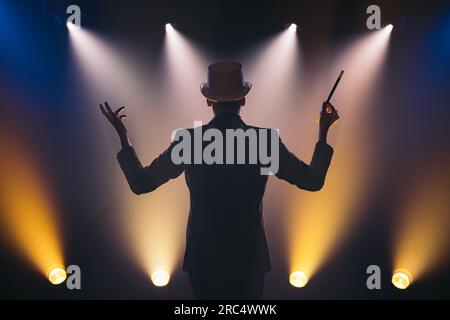 Vue arrière d'illusionniste masculin en costume et chapeau haut de gamme avec bâton magique faisant la présentation sur scène sombre dans le cirque Banque D'Images