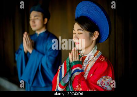 Portrait de jeune couple asiatique en tenue tendance et chapeau priant avec les mains serrées et les yeux fermés tout en se tenant dans la chambre sombre à Hue Vietnam Banque D'Images