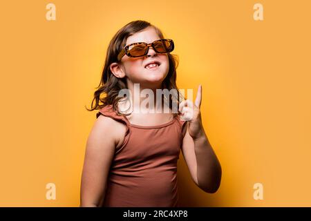 Drôle petite fille joyeuse avec des cheveux bruns ondulés dans des lunettes de soleil à la mode et maillot de bain souriant avec les yeux fermés et pointant vers le haut sur fond orange i. Banque D'Images