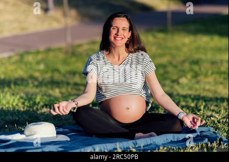 Corps entier de yogi femelle enceinte avec le gros ventre méditant avec les mains dans le mudra de Gyan sur la pelouse verte Banque D'Images