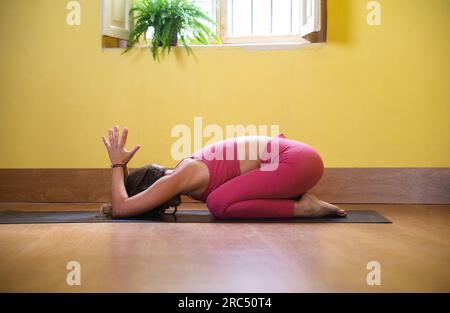 Vue latérale de jeune femme flexible méconnaissable en vêtements de sport assis sur le tapis dans Balasana yoga pose avec les mains levées dans Anjali Mudra pendant la pratique Banque D'Images