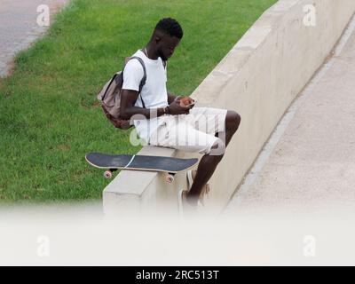 Angle élevé de l'homme afro-américain avec sac à dos assis sur le trottoir en ville avec skateboard près et en utilisant smartphone Banque D'Images
