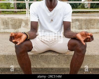 Crop méconnaissable afro-américain masculin millennial en été tenue décontractée faisant un geste mudra tout en étant assis sur des marches de pierre et en méditant dans le parc Banque D'Images