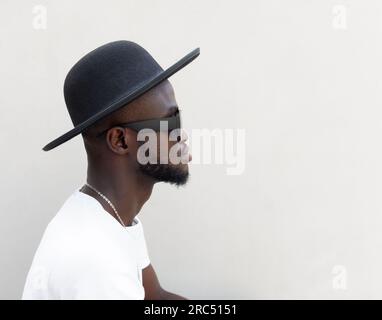 Vue de côté de jeune barbu noir en tenue tendance lunettes de soleil et chapeau assis sur des escaliers en pierre près du mur blanc et regarder loin sur la rue Banque D'Images