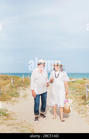 Heureux homme âgé et femme en vêtements de plage et chapeaux marchant sur le chemin de la plage sous la lumière du soleil Banque D'Images