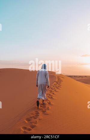 Vue arrière d'un homme berbère méconnaissable en vêtements traditionnels marchant sur une dune de sable dans le désert de Merzouga au coucher du soleil Banque D'Images