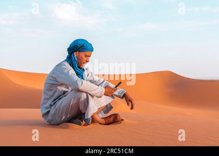 Vue latérale de l'homme berbère dans les vêtements traditionnels touaregs assis sur le sable tout en naviguant smartphone contre le ciel bleu dans le désert de Merzouga Maroc Banque D'Images