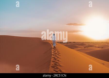 Vue arrière lointaine de l'homme berbère méconnaissable en vêtements traditionnels marchant sur une dune de sable dans le désert de Merzouga au coucher du soleil Banque D'Images