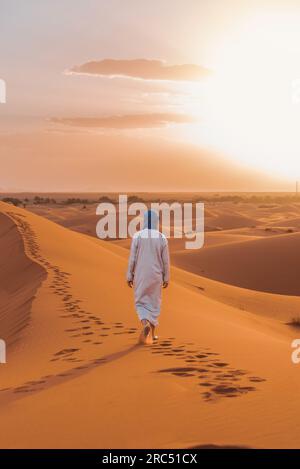 Vue arrière d'un homme berbère méconnaissable en vêtements traditionnels marchant sur une dune de sable dans le désert de Merzouga au coucher du soleil Banque D'Images