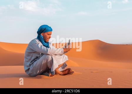 Vue latérale de l'homme berbère dans les vêtements traditionnels touaregs assis sur le sable tout en naviguant smartphone contre le ciel bleu dans le désert de Merzouga Maroc Banque D'Images