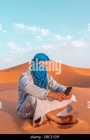 Corps entier de l'homme berbère dans les vêtements traditionnels touareg assis sur le sable tout en naviguant smartphone contre le ciel bleu dans le désert de Merzouga Maroc Banque D'Images