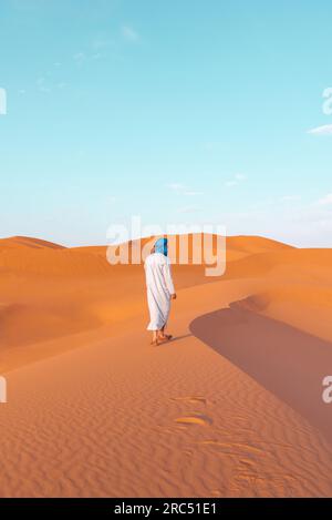 Vue arrière du corps entier de l'homme berbère anonyme portant des vêtements traditionnels touareg et des lunettes de soleil marchant sur la dune de sable tout en regardant loin dans Merzouga d Banque D'Images