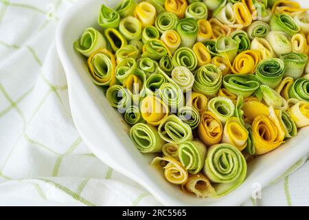 Petits rouleaux de courgettes vert et jaune avec sauce crémeuse dans un plat blanc allant au four sur fond d'une serviette à carreaux Banque D'Images