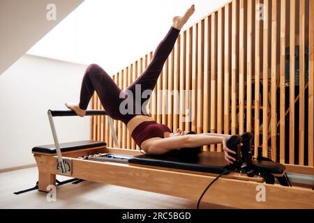 Une jeune fille fait du Pilates dans un studio lumineux sur un lit reformeur. Mince brunette en body bordeaux fait des exercices pour renforcer les muscles du dos, Banque D'Images