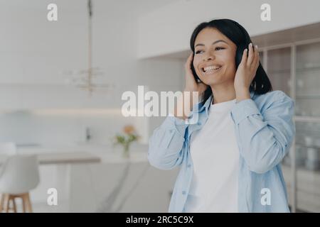 Femme joyeuse à la maison écoute de la musique ou des podcasts avec des écouteurs sans fil modernes. Souriante, elle apprécie ses chansons préférées et ses livres audio avec Perfect Banque D'Images