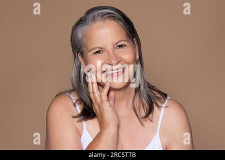 Joyeuse dame caucasienne âgée avec les cheveux gris touche le visage avec la main, apprécie le traitement anti-âge Banque D'Images