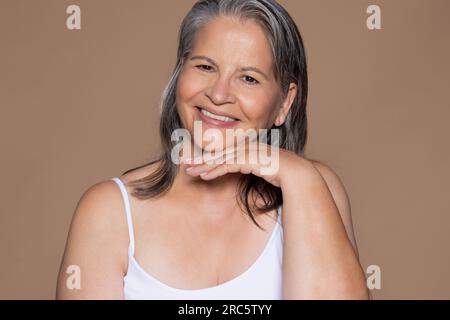 Heureuse femme caucasienne âgée avec les cheveux gris touche le visage avec la main, apprécie le traitement anti-âge Banque D'Images